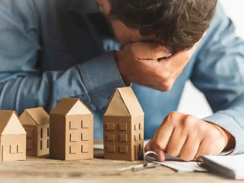Propietario preocupado con la cabeza entre las manos junto a miniaturas de edificios, representando la angustia de un inquilino que deja de pagar el alquiler